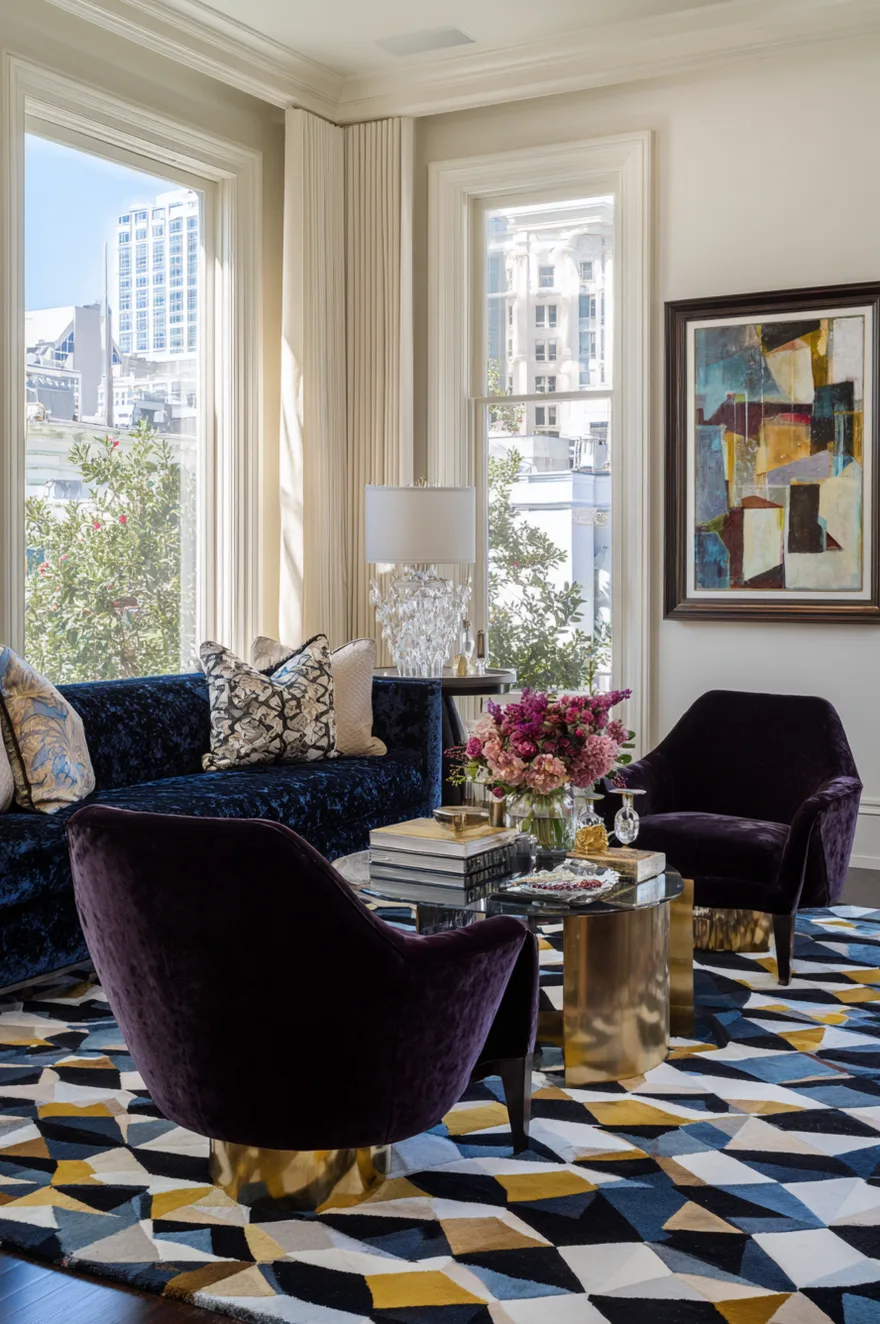 Sunlit living room featuring a large geometric rug with blue, gold, and black triangle patterns, a deep blue velvet sofa, two plum velvet accent chairs with brass cylinder bases, and a gold coffee table.