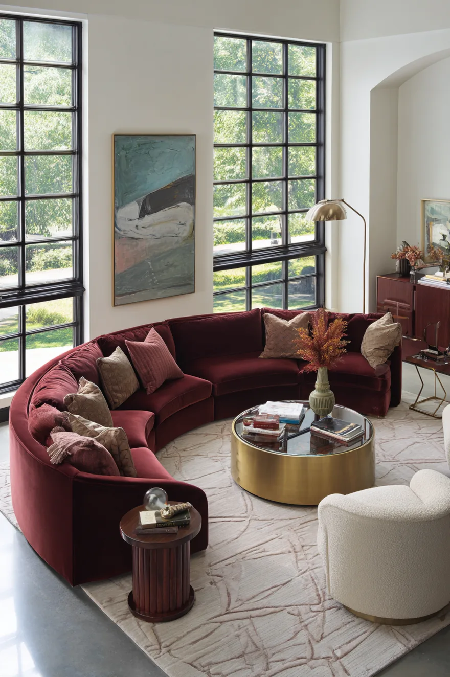 High-ceiling living room featuring a large curved burgundy velvet sectional sofa, a round glass-top coffee table with a brass cylinder drum base, and a white bouclé armchair.
