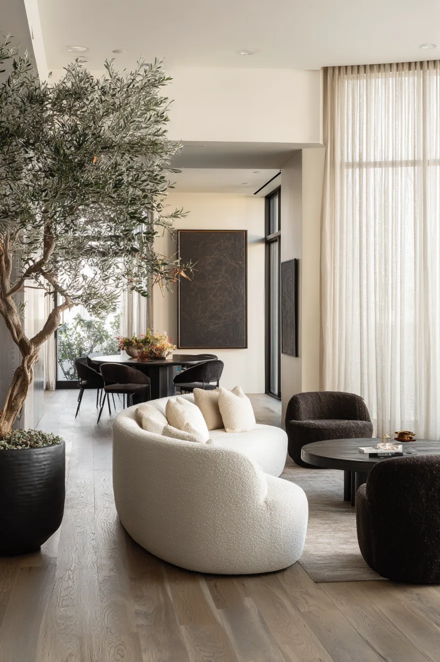 A bright open plan room featuring a curved white boucle sofa paired with two dark brown club chairs around a black coffee table, leading to a dark round dining table in the background.