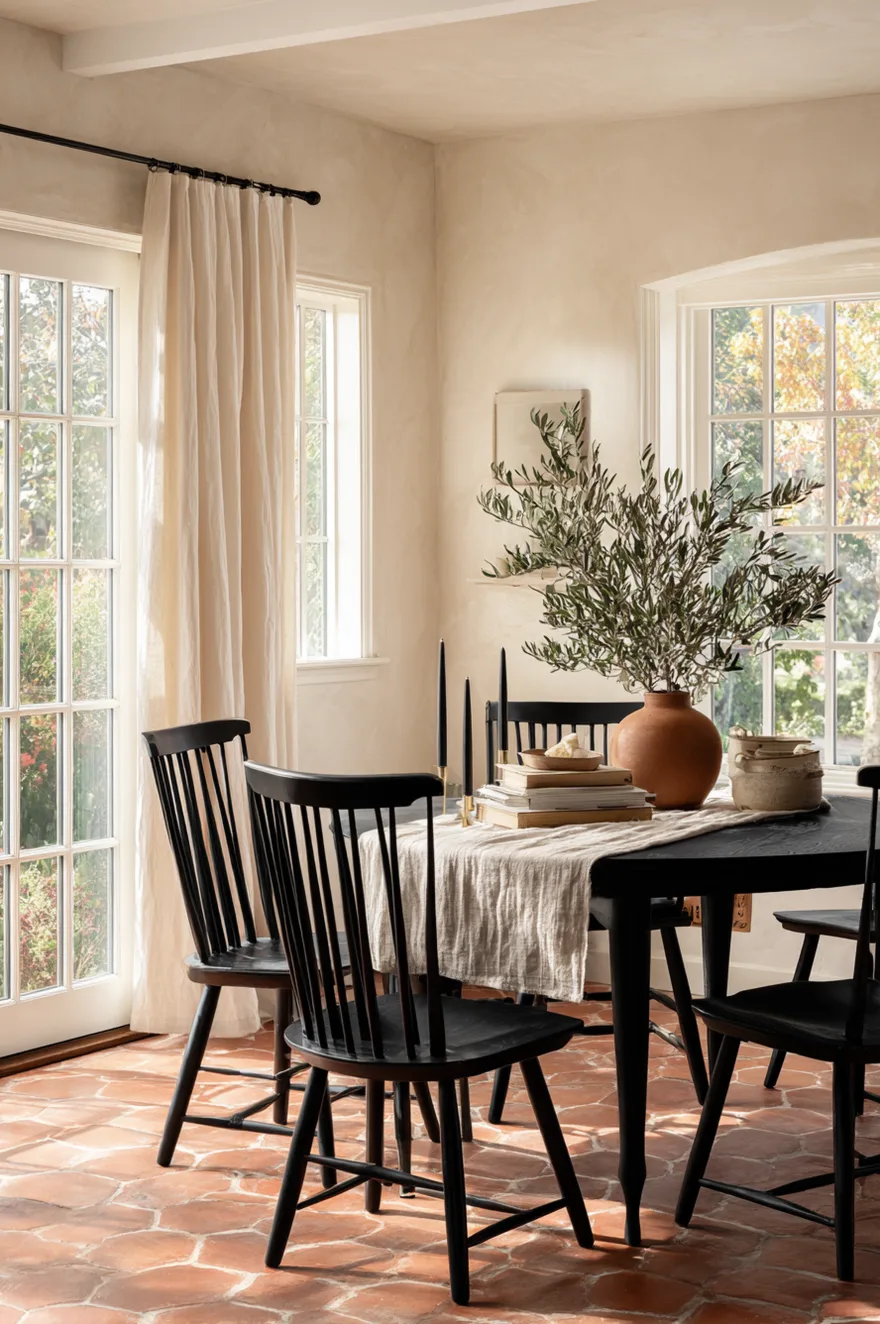 Windsor spindle-back chairs in distressed black finish around a round dining table with terracotta hex tile floor, cream plaster walls, and brass taper candle holders.