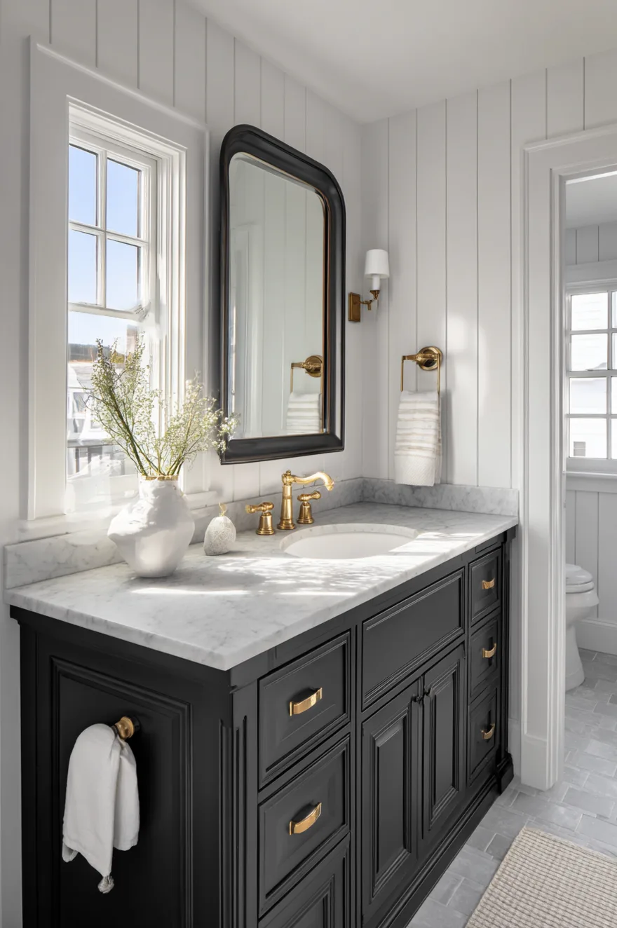 Dark near-black inset panel vanity cabinet with a honed Carrara marble countertop, white undermount sink, unlacquered brass faucet, beveled mirror, and white shiplap upper walls in a bright farmhouse guest bathroom.