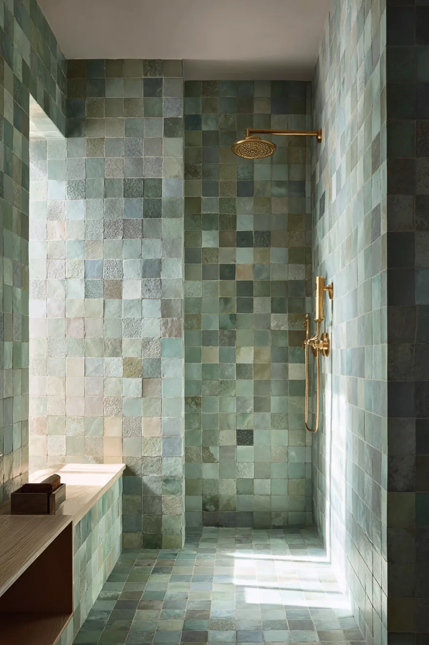 A walk in shower completely covered in square handmade tiles in varying shades of soft green and blue featuring a wall mounted brass shower head and controls.